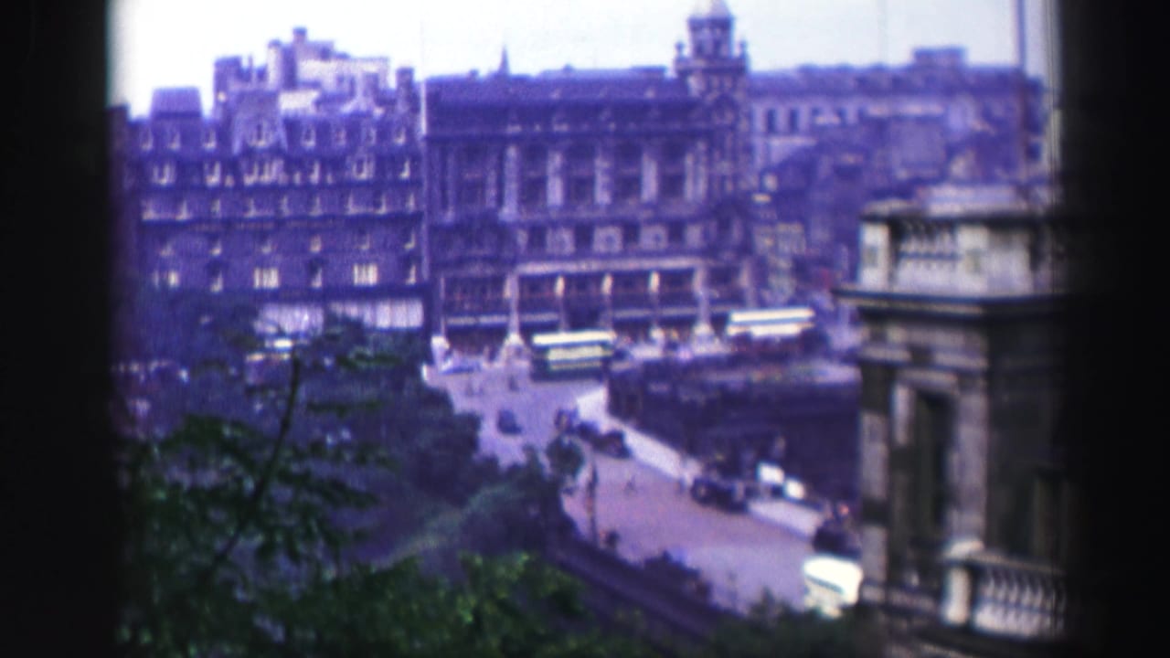 High-angle city street seen through a window with stone buildings, buses, cars, and trees.