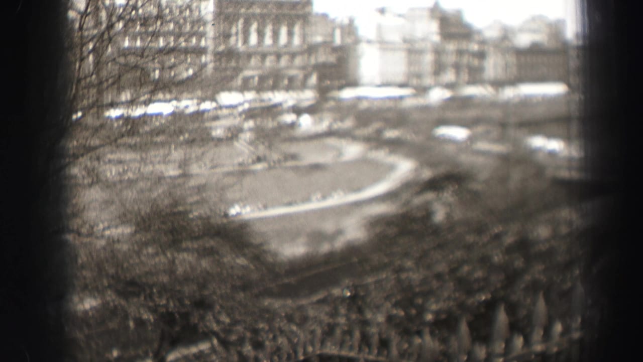 Soft 8mm frame of an Edinburgh park overlook, showing bare branches, a curved roadway, small vehicles, awnings, and stone facades.