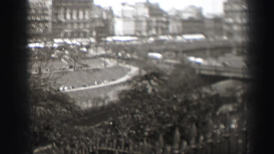1952 Edinburgh 8mm frame with a curved park road, passing cars, foreground branches and railings, and distant urban buildings.
