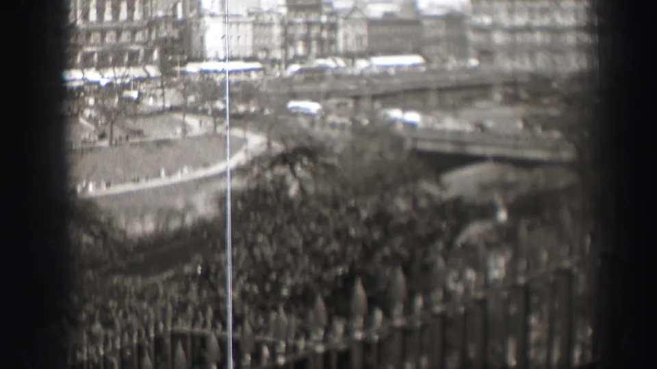 Final 8mm frame from an Edinburgh park overlook, with cars near a bridge or raised roadway, railings, bare trees, and city buildings.
