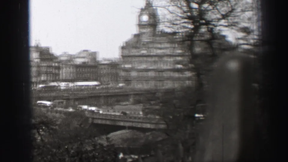 1952 Edinburgh home-movie view with a clock tower above bridge traffic, seen past trees and a blurred foreground railing.