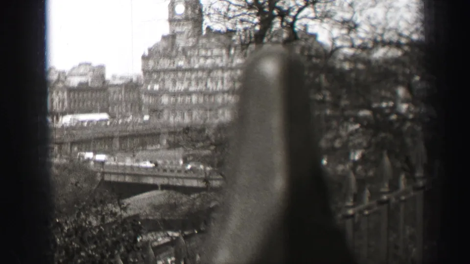 1952 Edinburgh home-movie frame looking past out-of-focus iron railings toward stone buildings, bare trees, and a city bridge.