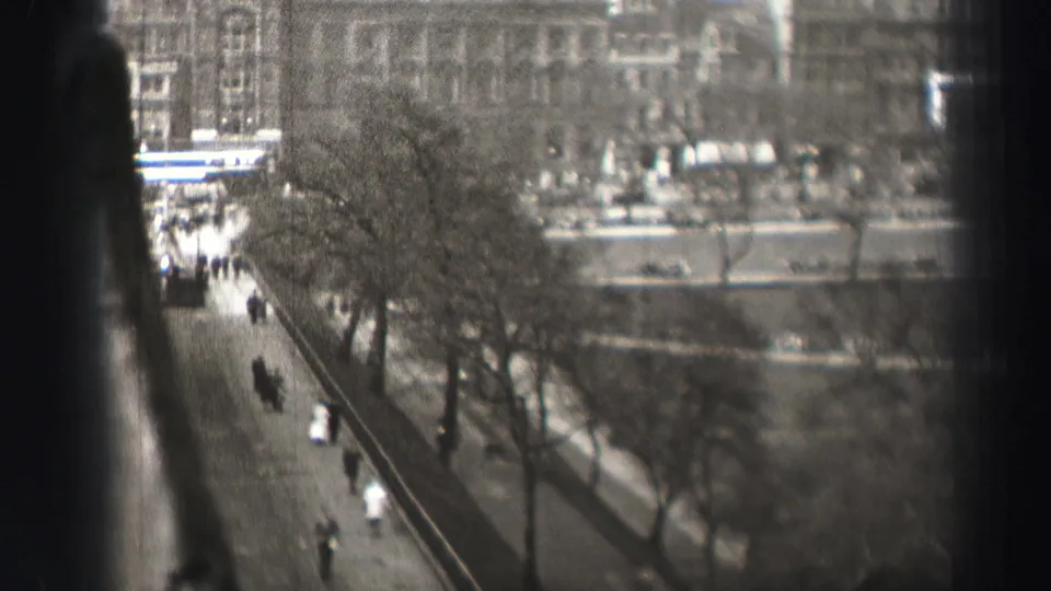 1952 Edinburgh frame looking down past railings to a tree-lined sidewalk, scattered pedestrians, roadway, bridge area, and background buildings.