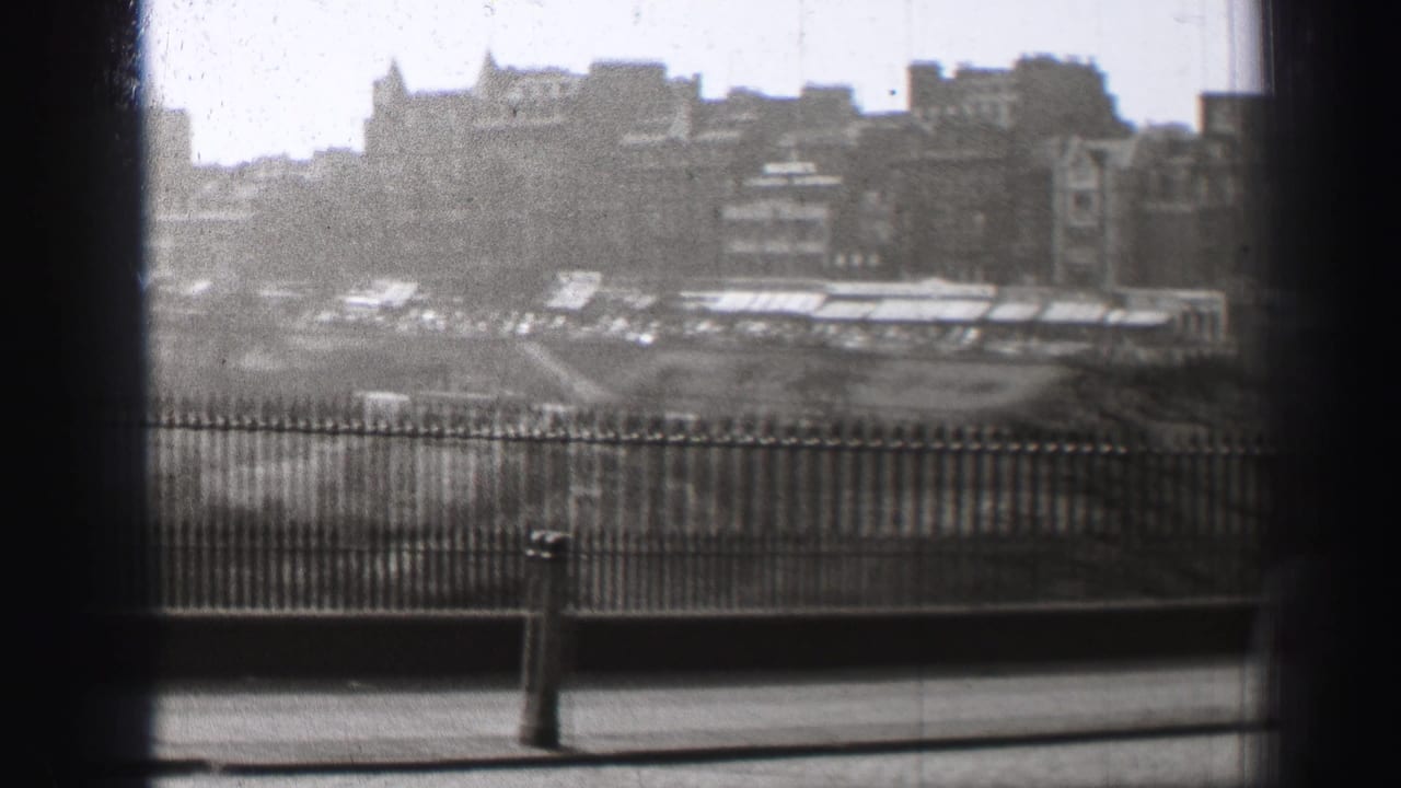 Black-and-white Edinburgh view with iron railings, a pavement edge, and dense buildings in the distance.