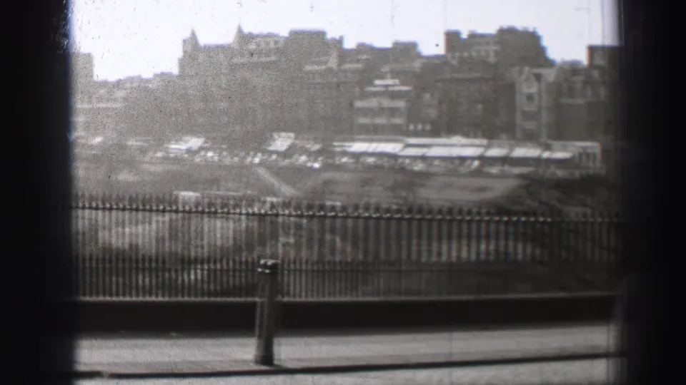 Black-and-white Edinburgh view with iron railings, a pavement edge, and dense buildings in the distance.