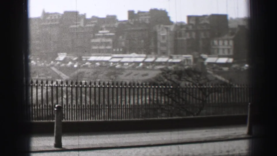 1952 Edinburgh street overlook with railings, awnings, clustered buildings, and small distant pedestrians or vehicles.