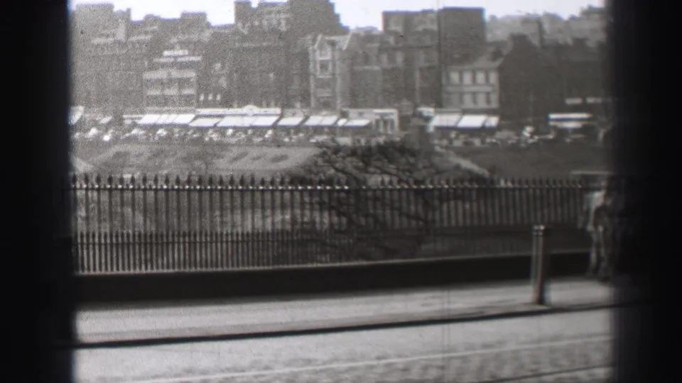 Grainy black-and-white 8mm frame of Edinburgh railings, a street post, distant awnings, and older city buildings.