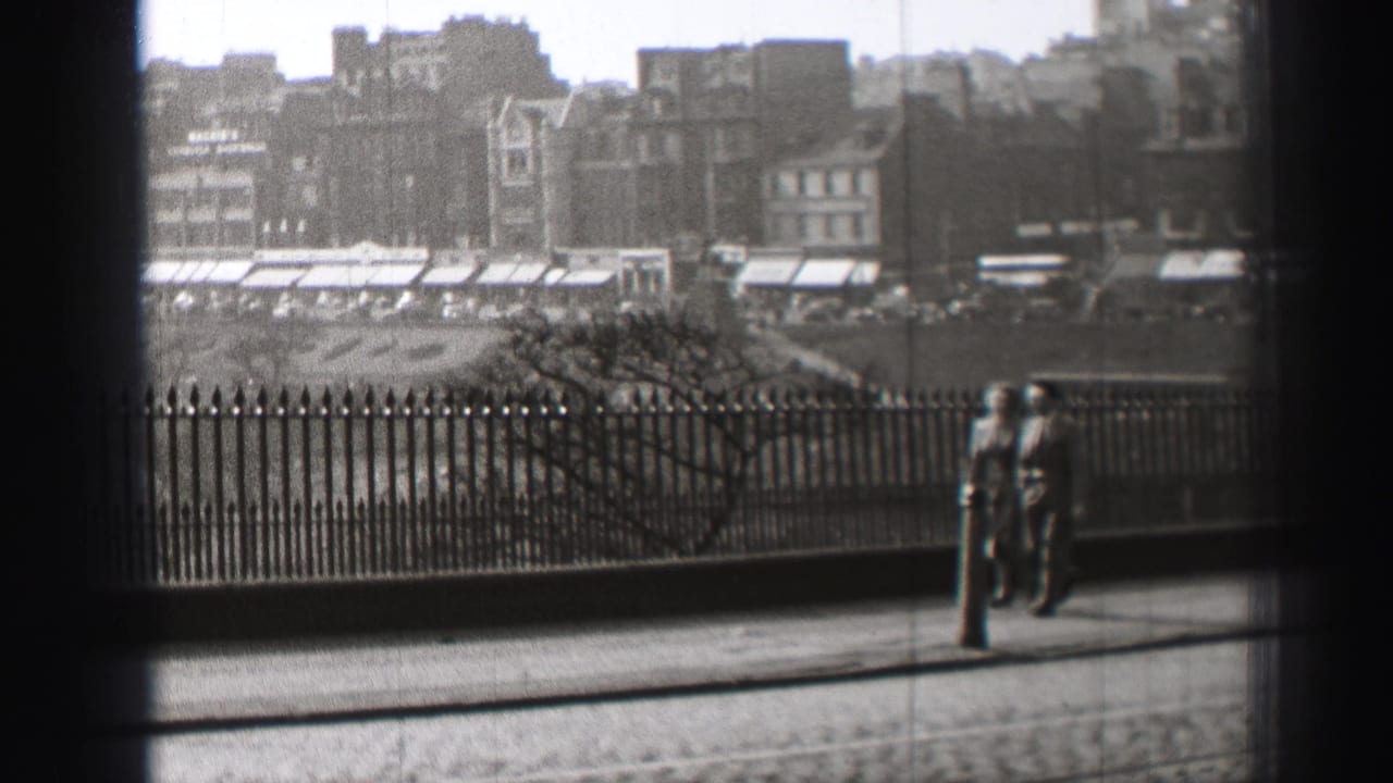 Pedestrians walk beside iron railings on a city street with buildings in the background.