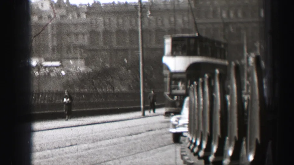 1952 Edinburgh street view with a double-decker trolley, pedestrians, older cars, and large stone buildings seen through a dark foreground barrier.