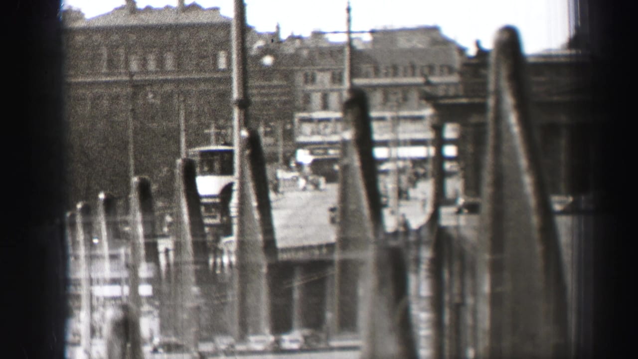 1952 Edinburgh home-movie frame looking through a dark window edge toward stone buildings, street traffic, poles, and soft foreground posts.