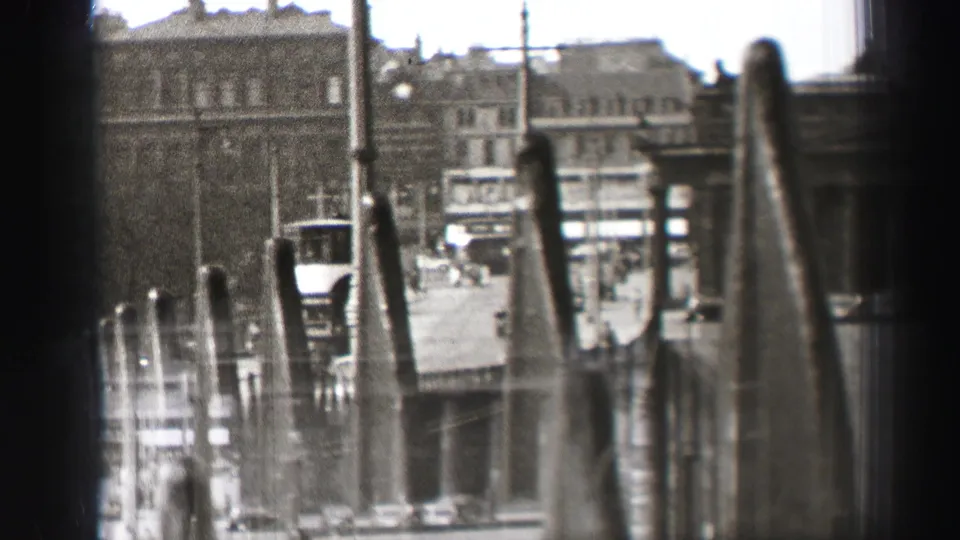 1952 Edinburgh home-movie frame looking through a dark window edge toward stone buildings, street traffic, poles, and soft foreground posts.