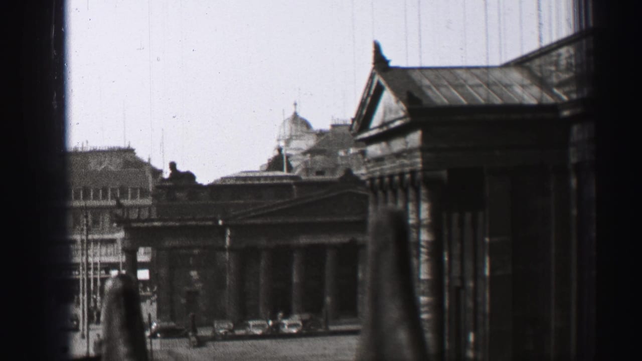 Columned stone building and nearby rooflines seen beyond dark window edges and blurred foreground posts.