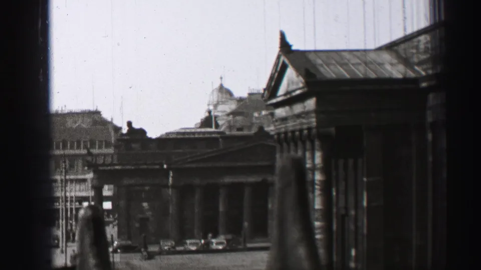 1952 Edinburgh view through a window, with a columned stone facade, rooflines, small cars, and indistinct figures in the distance.