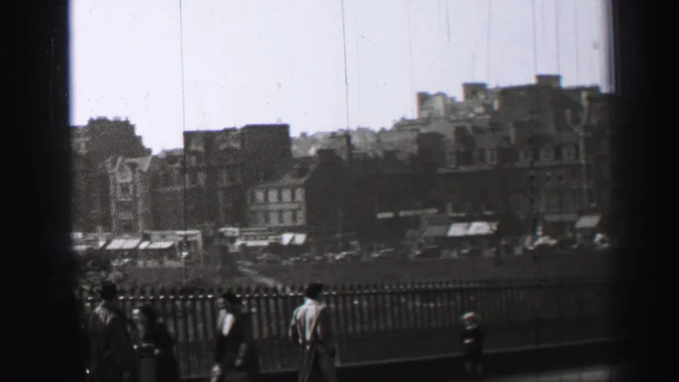 Black-and-white Edinburgh frame with several pedestrians beside an iron railing, older buildings beyond, and faint vehicles in the background.