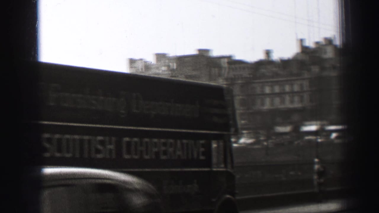 1952 Edinburgh street view from a moving vehicle, with a double-decker bus, car roof, bridge railing, and stone buildings beyond.