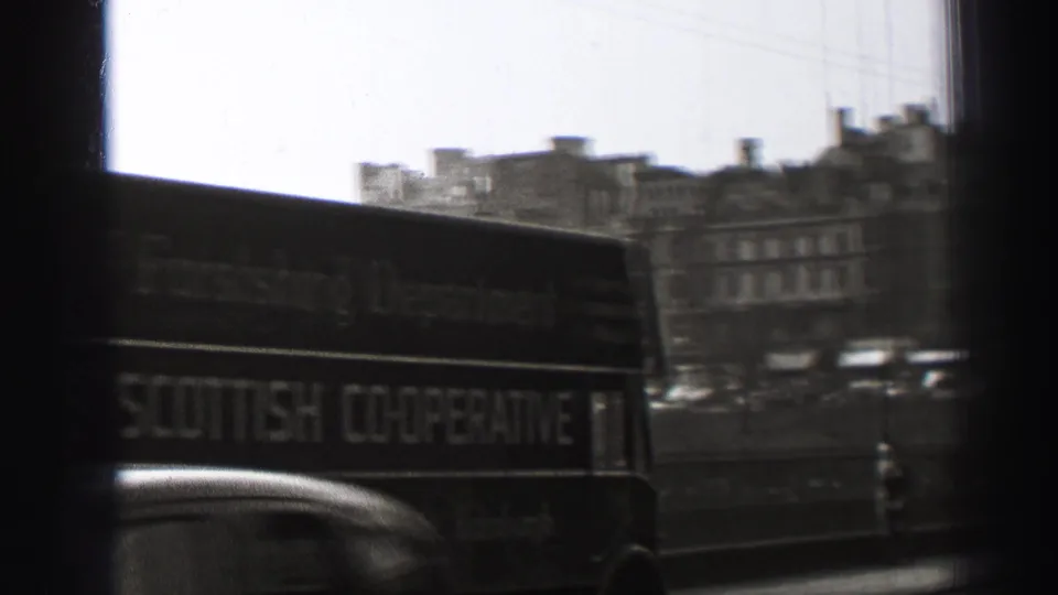 1952 Edinburgh street view from a moving vehicle, with a double-decker bus, car roof, bridge railing, and stone buildings beyond.