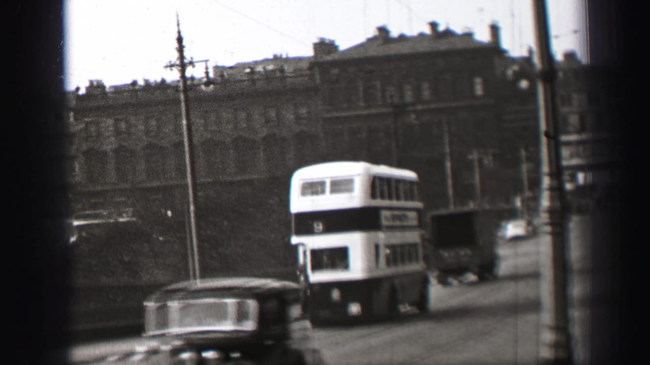 Double-decker bus farther down the road with a car, truck, railings, and buildings nearby.
