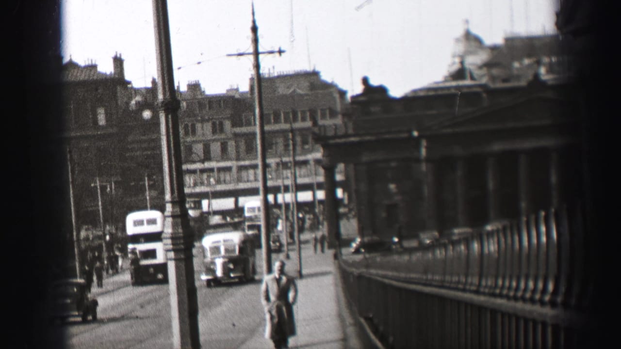 Pedestrian in a long coat beside traffic, railings, poles and city buildings.