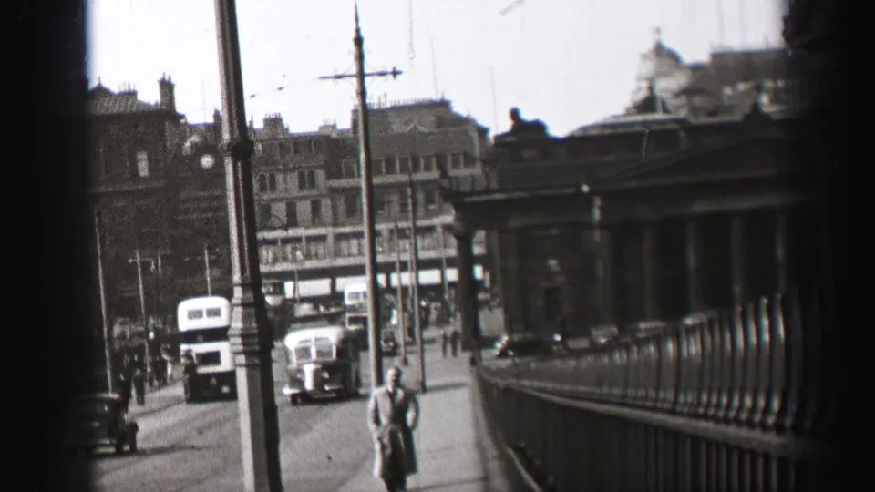 1952 Edinburgh sidewalk scene with a pedestrian in a long coat, bus, car, railings, street poles and buildings.