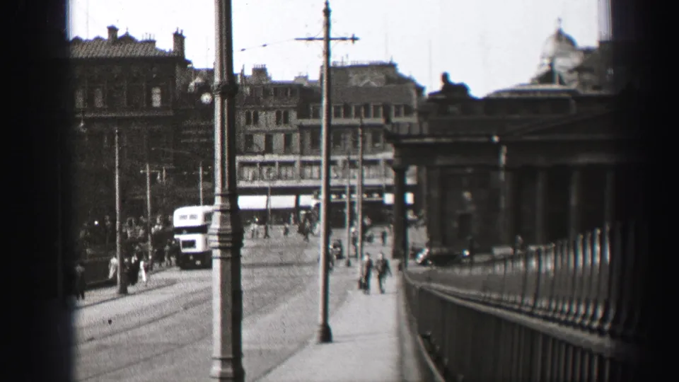 1952 Edinburgh sidewalk and road view with pedestrians, a bus, railings, lampposts, and layered city buildings.