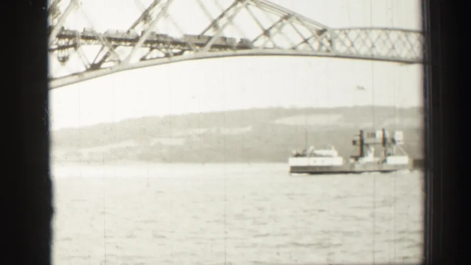 Soft 8mm view of a ferry or work vessel beneath a steel bridge span near Edinburgh, with shoreline hills in the distance.