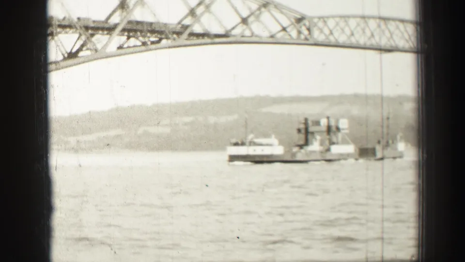 1952 home-movie frame showing a ferry or workboat near a smaller craft under the steel bridge, with water and hills behind.