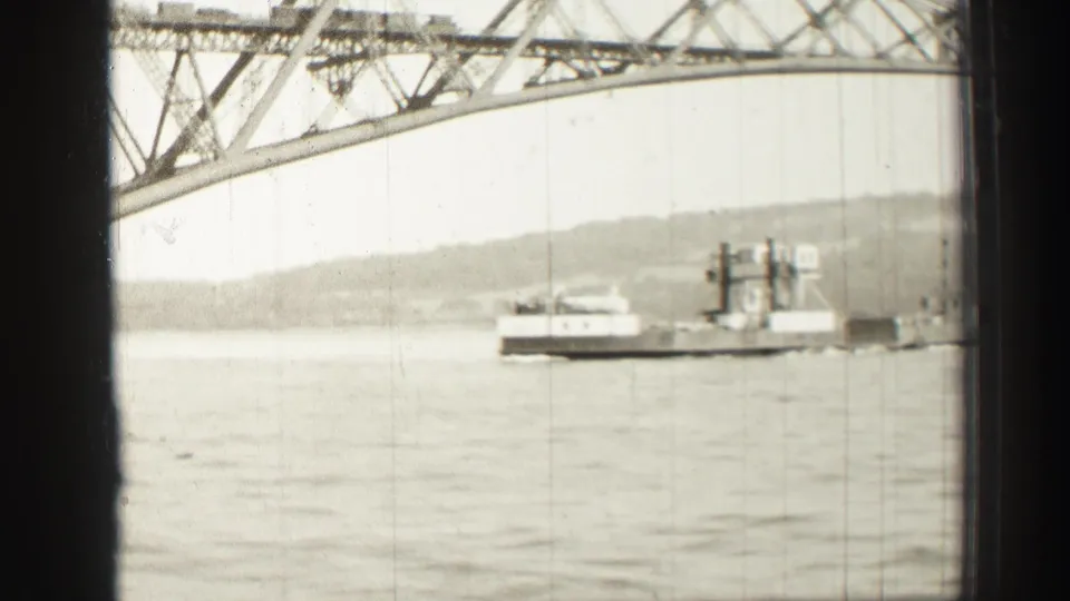 Black-and-white 8mm view of a ferry crossing the river below a large steel truss bridge in Scotland.