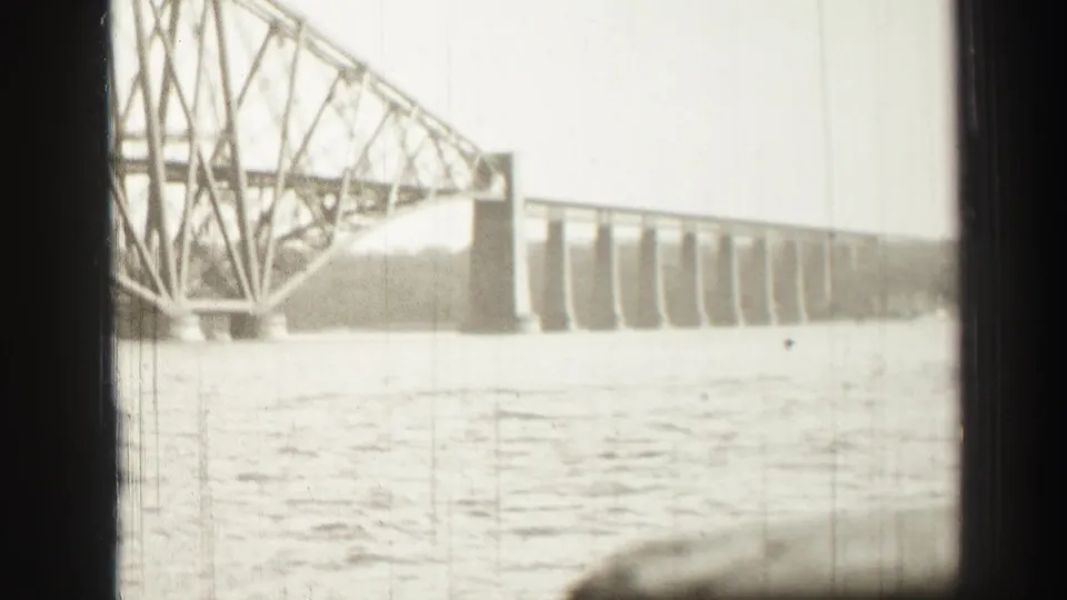 8mm black-and-white frame of a large steel bridge over water, with shoreline visible beyond the span.