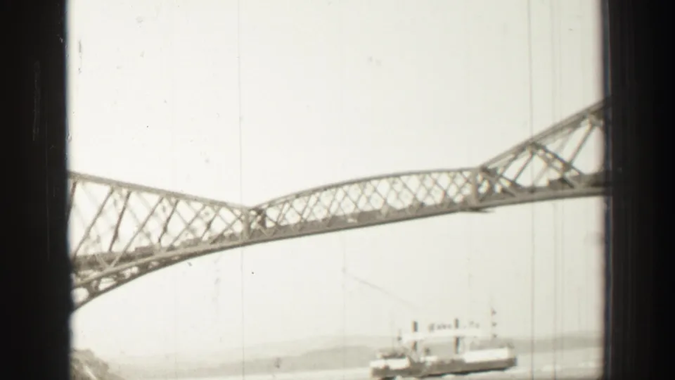 1952 8mm travel footage near Edinburgh showing the bridge span sweeping across the frame with a ship below and shoreline in the distance.