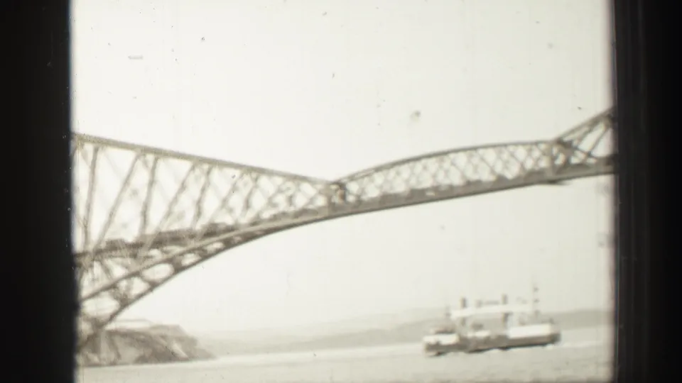 1952 home-movie frame near Edinburgh with a steel truss bridge crossing above water as a commercial ship moves near the lower edge.