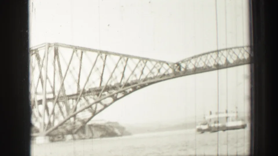 The Forth Bridge appears through dark boat-window bars, showing the steel cantilever structure, water, shoreline, and a small vessel.
