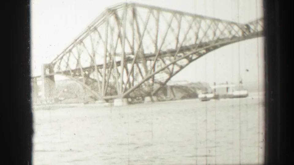 The bridge remains centered from the moving boat, with its cantilever trusswork, water, shoreline, and distant vessel visible through film wear.