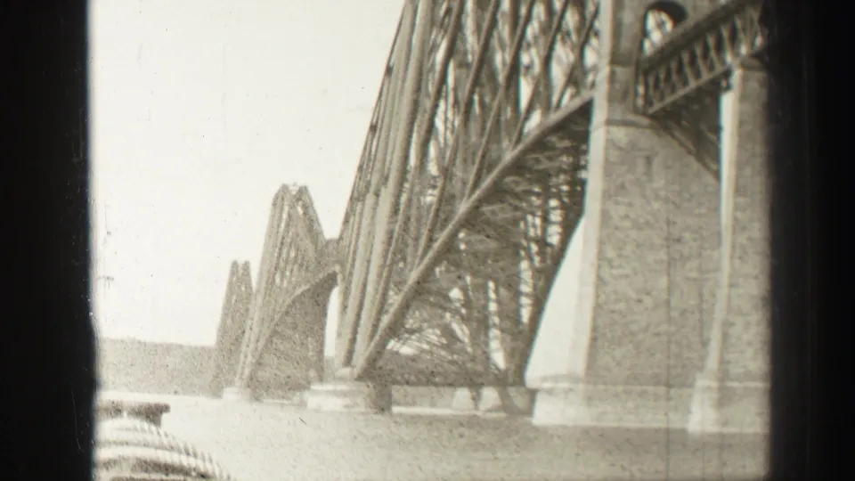 1952 home-movie frame looking along the Forth Bridge supports, with repeating steel spans and masonry piers over the water.