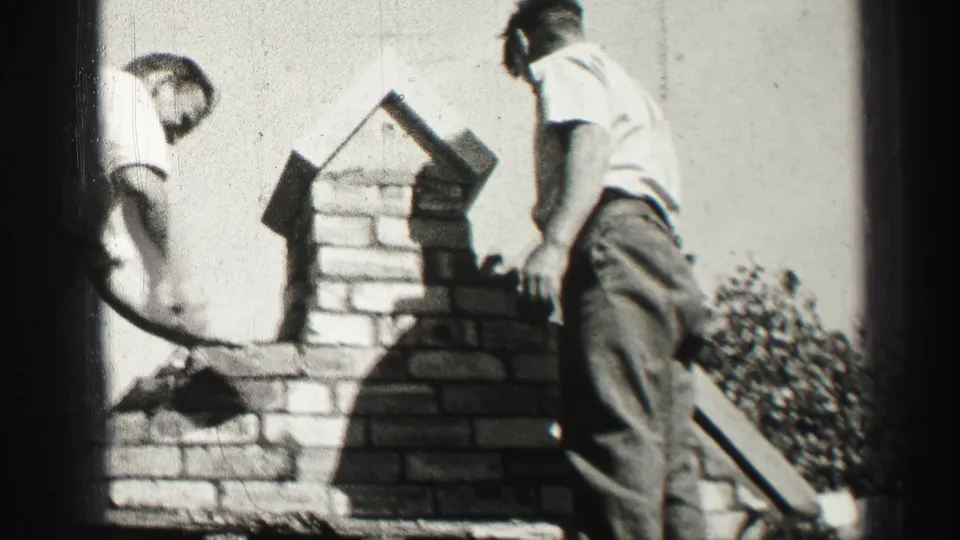 1952 8mm frame of two men beside brickwork, one bending near the wall and another standing with a tool outdoors.