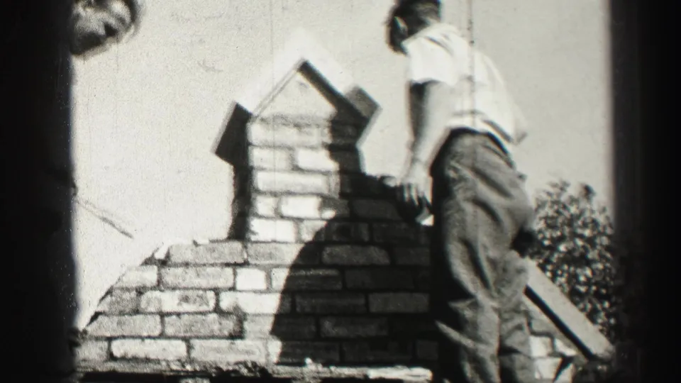 1952 Edinburgh home-movie frame showing a man beside a partly built brick chimney on a roof in bright daylight.