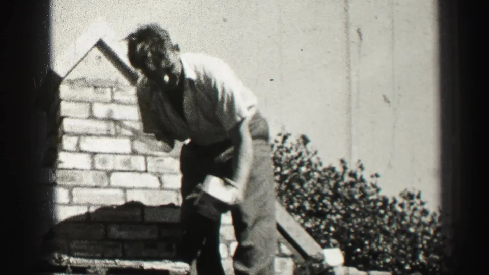 Black-and-white 8mm frame of a man bending beside a brick chimney during rooftop work in Edinburgh, Scotland.