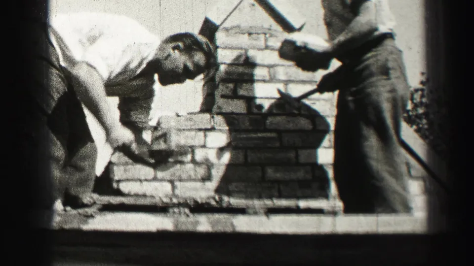 Two adult men work with mortar and bricks on a small masonry structure in 1952 Edinburgh home-movie footage.