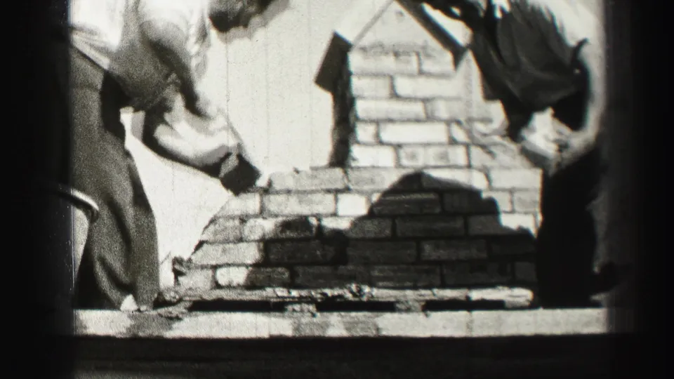A close home-movie view shows one man applying mortar to brickwork while another worker stands nearby in Edinburgh.
