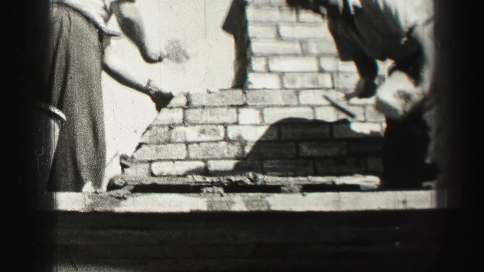 Two adult men lean over a partially built brick structure, continuing hand masonry work in black-and-white 8mm footage.