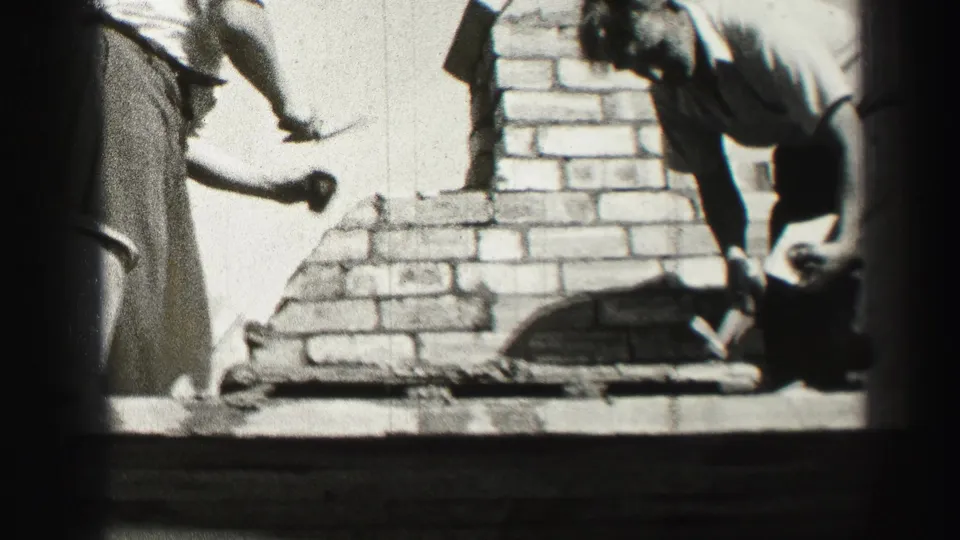 At the end of the clip, one worker bends beside the brickwork as another reaches in with a masonry tool.