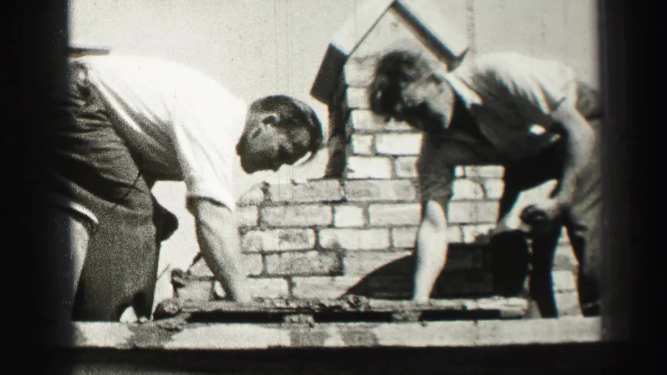 In 1952 Scotland home-movie footage, two adult workers bend over brick courses while building or repairing a chimney with trowels.