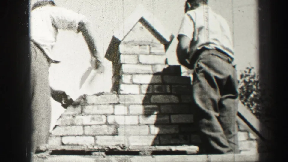 A 1950s rooftop masonry view shows two adult workers beside a partially built chimney, with one reaching toward fresh mortar.