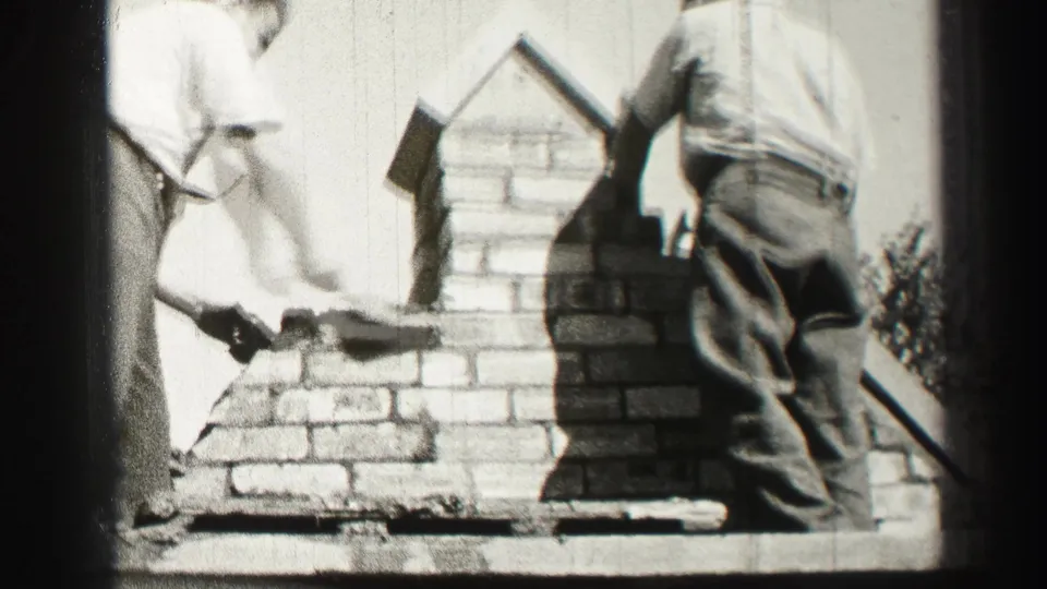 Black-and-white 8mm footage shows an adult worker troweling mortar on a rooftop chimney while another stands close by.