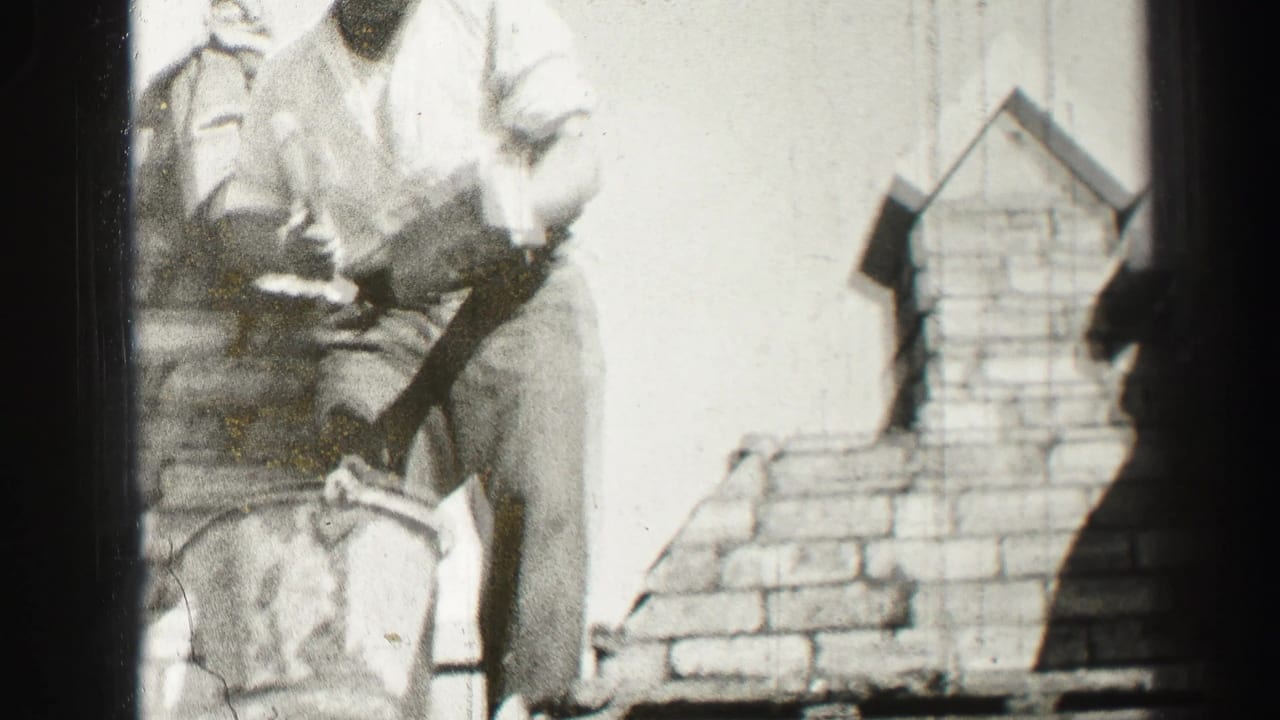 Black-and-white 1952 home-movie frame from Edinburgh showing an adult bricklayer leaning over a partially built brick chimney or wall section.