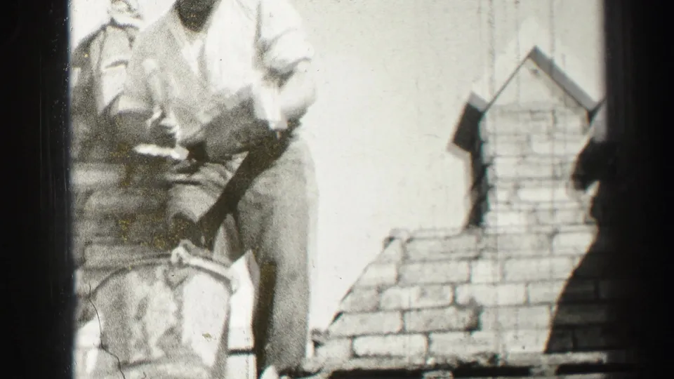 Black-and-white 1952 home-movie frame from Edinburgh showing an adult bricklayer leaning over a partially built brick chimney or wall section.