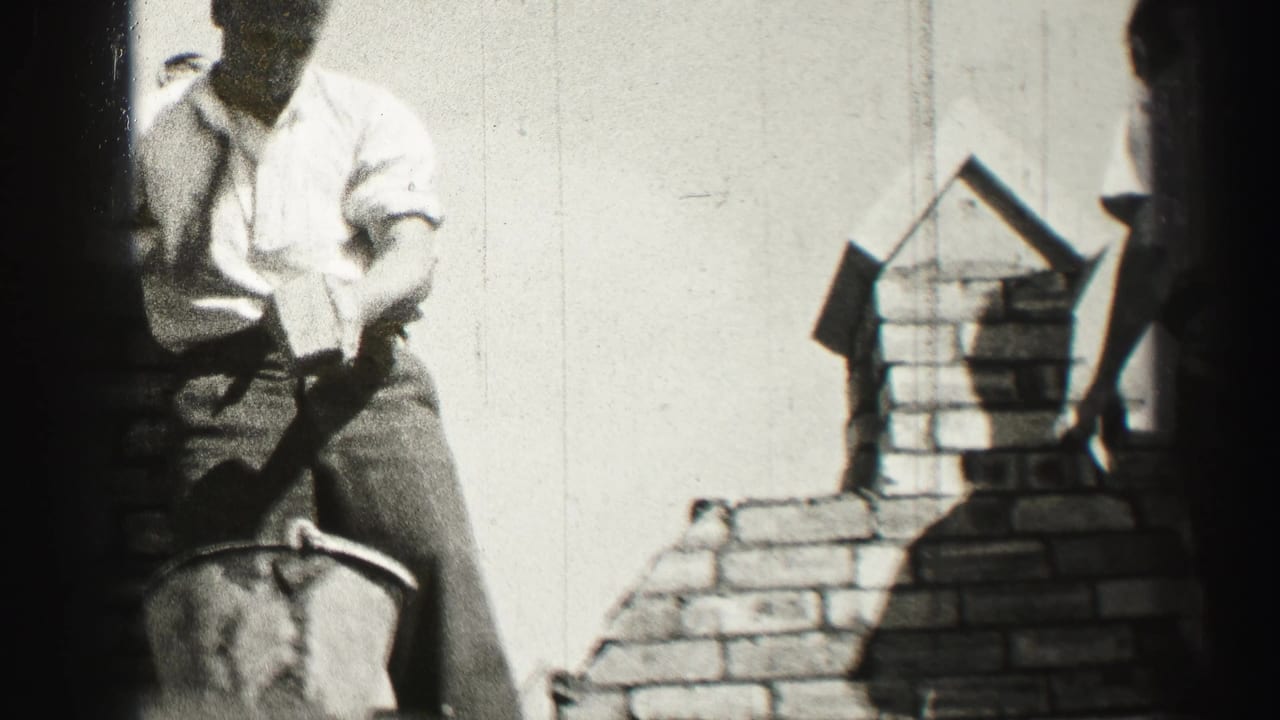 Black-and-white frame shows a bricklayer in shirtsleeves leaning over the brickwork, with the chimney outline and film edge visible.