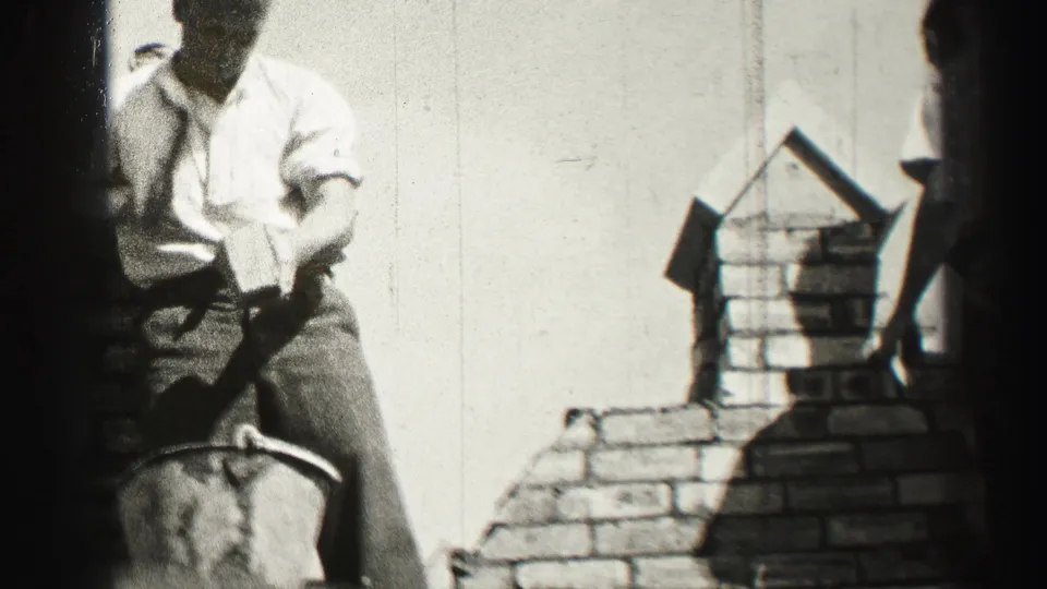 Black-and-white frame shows a bricklayer in shirtsleeves leaning over the brickwork, with the chimney outline and film edge visible.