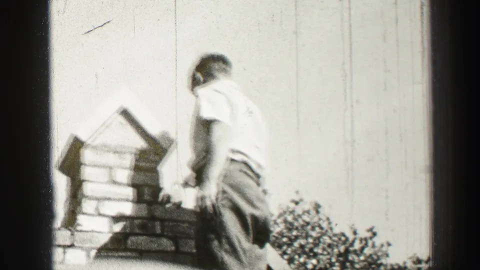 Black-and-white 8mm frame shows an adult bricklayer from behind, standing beside a partly built chimney with shrubs at the lower right.