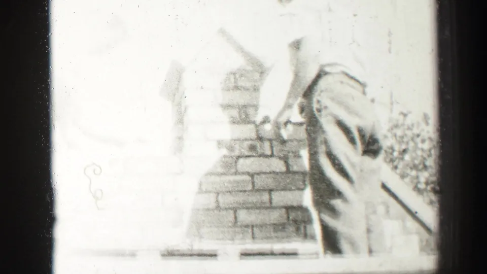 Overexposed black-and-white frame from the 1952 Edinburgh clip shows two adult men close to the brick chimney, with one figure partly cropped at the left.