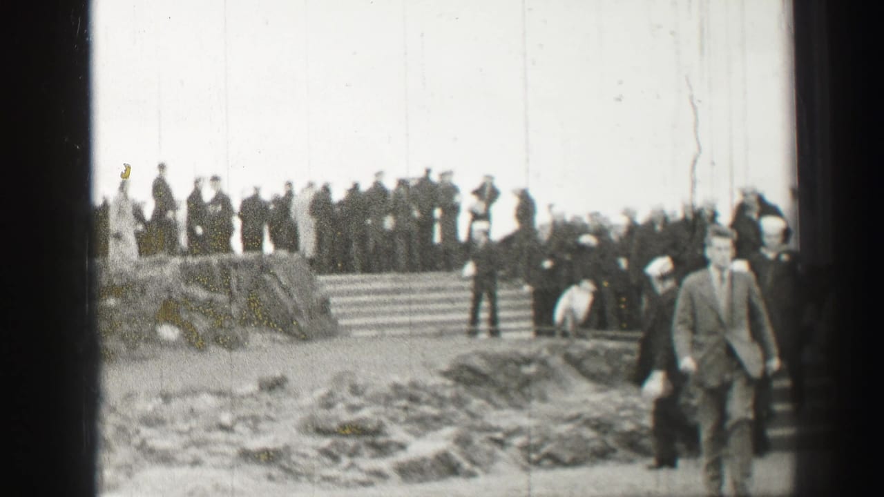 Crowd and uniformed men on stone steps as a suited man walks near the camera.